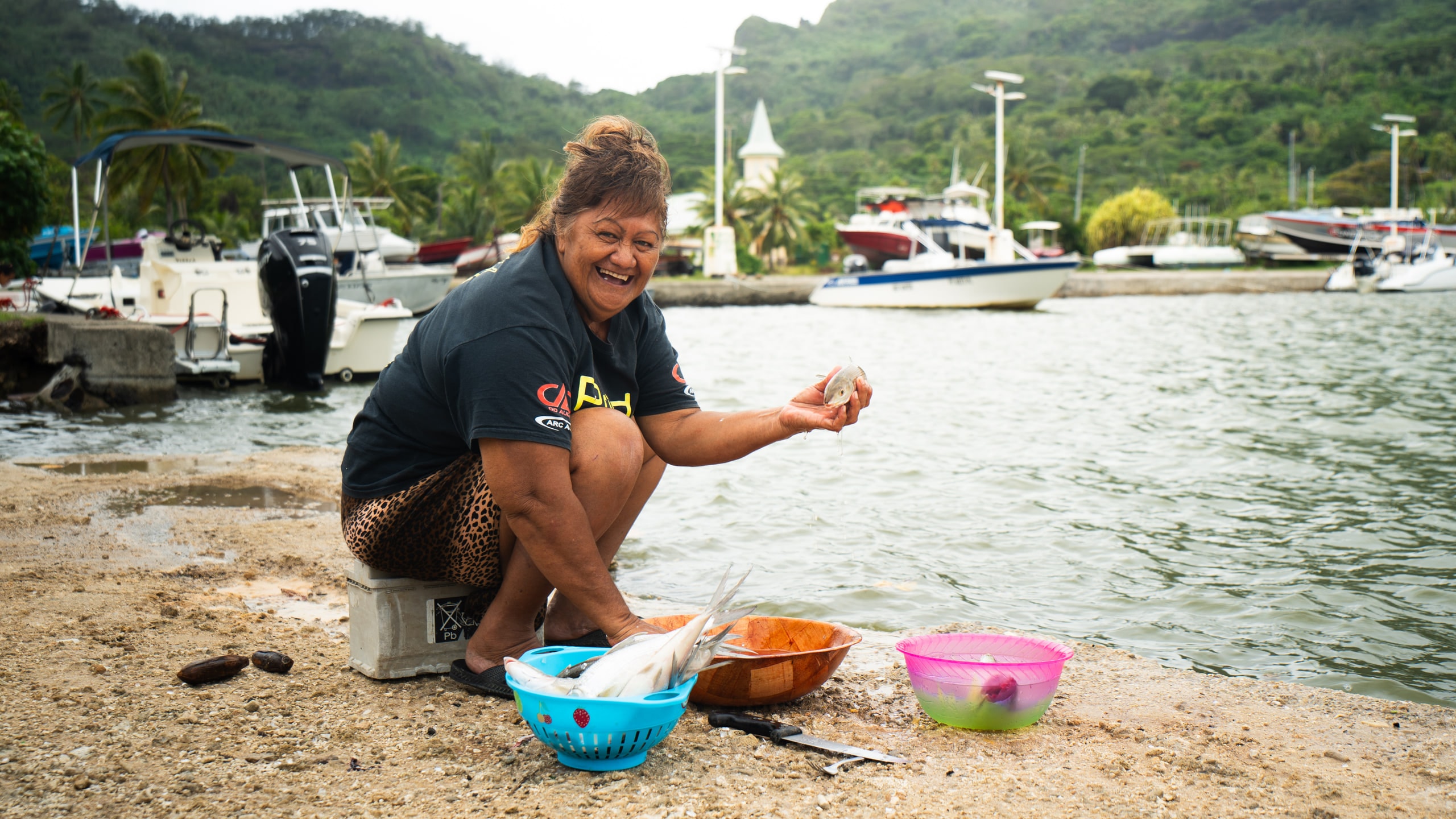 A woman preparing food at the water's edge beside moored boats
