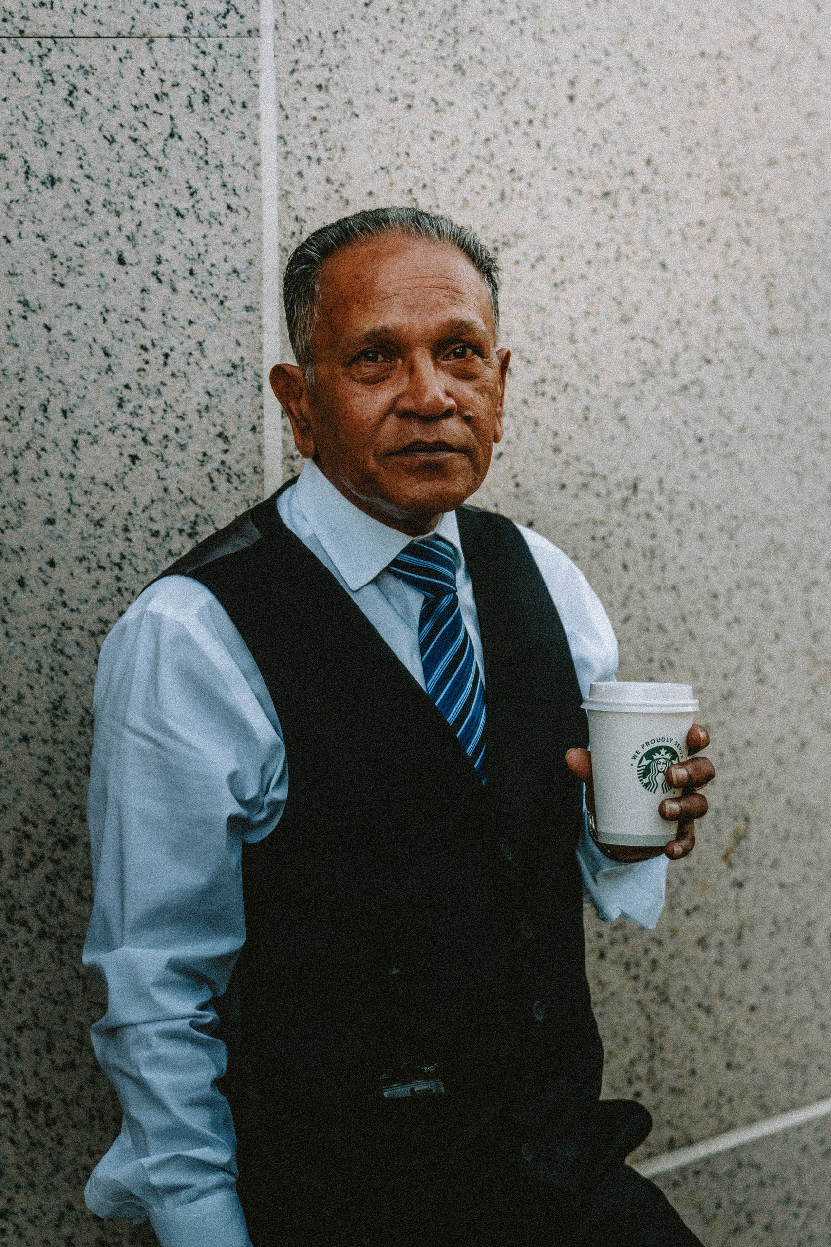 A man in a waistcoat and tie standing against a wall with a coffee cup